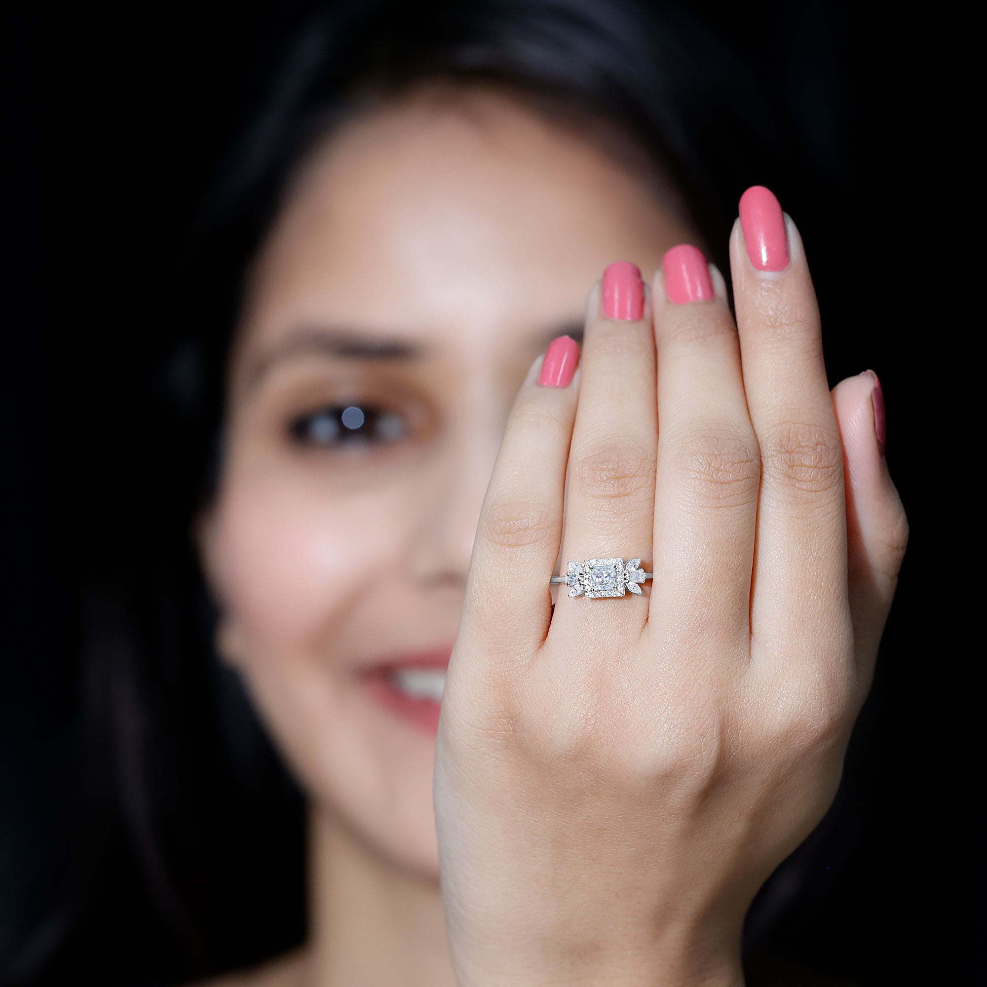 Woman wearing a white gold moissanite ring on her finger, with pink nail polish and a softly blurred smiling face in the background.