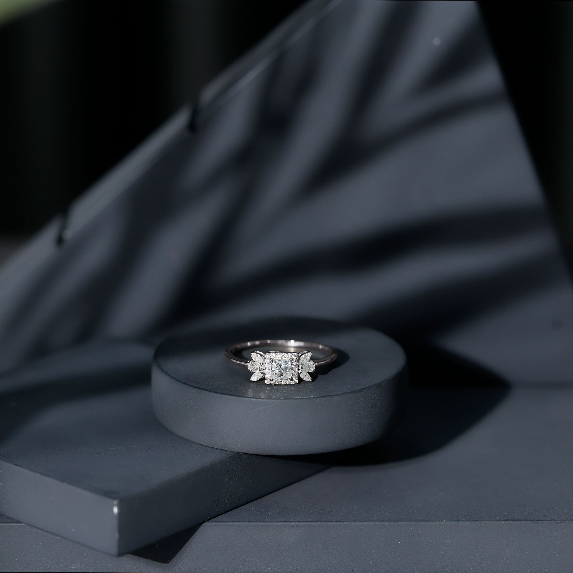 White gold princess cut moissanite ring displayed on a dark gray pedestal with a moody black fabric background, highlighting the bright center stone and floral side accents.
