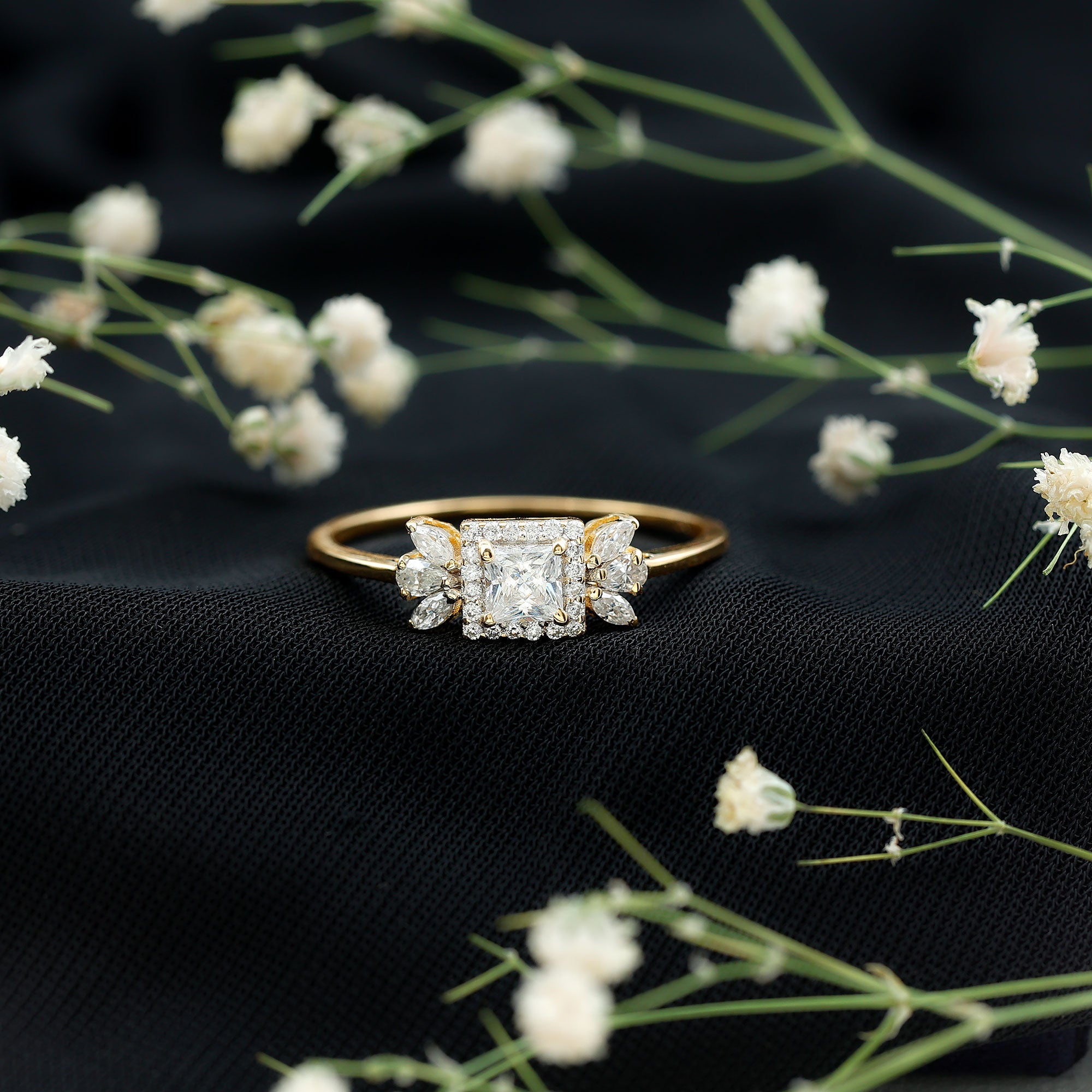 Yellow gold moissanite ring placed on black fabric with small white flowers, featuring a princess cut center stone, halo design, and floral side stone arrangement.