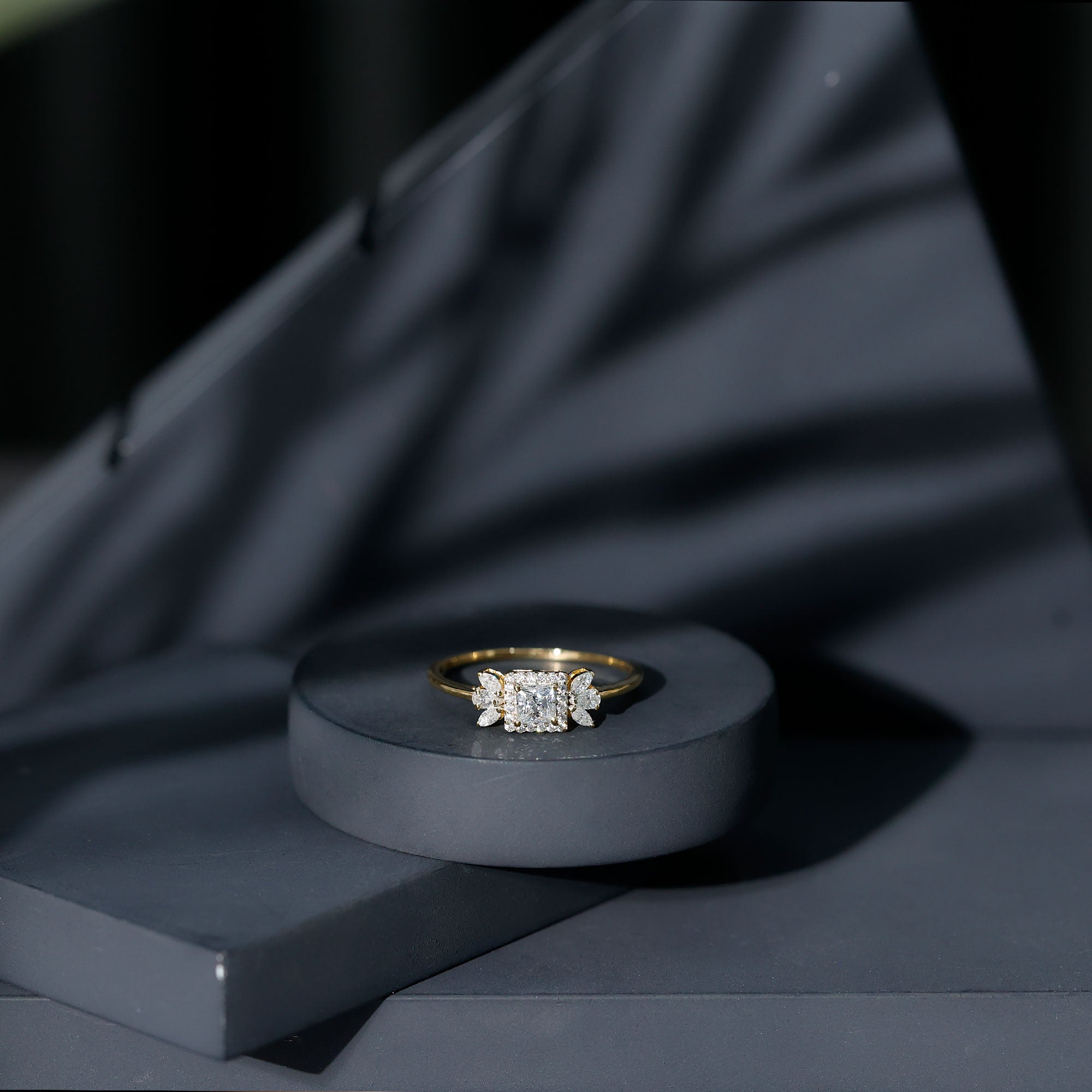 Yellow gold princess cut moissanite ring displayed on a dark gray pedestal with black draped fabric in the background, emphasizing the bright stones and warm gold band.