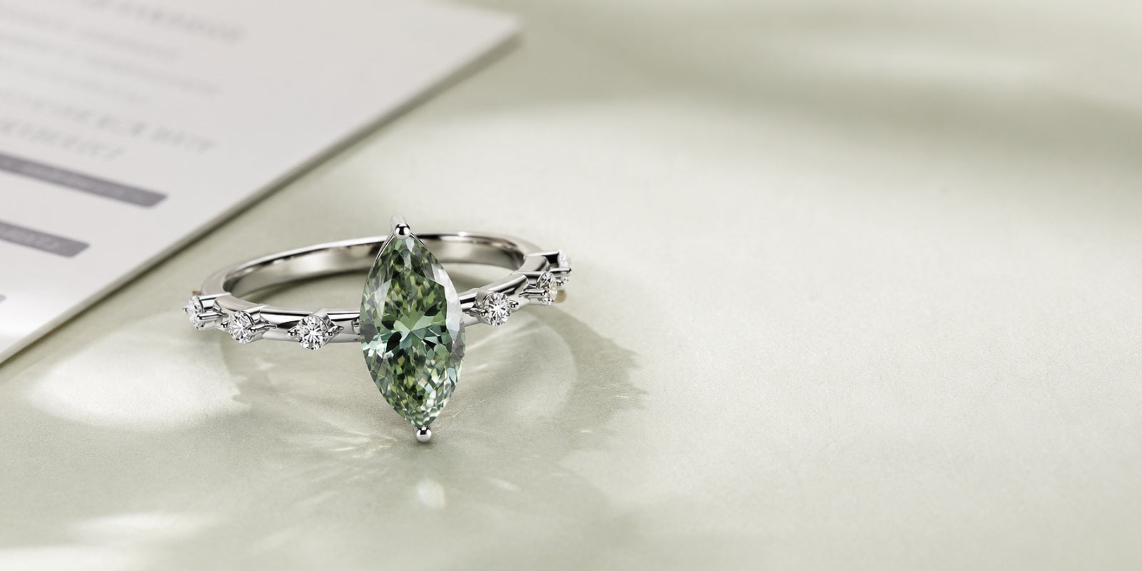 Marquise lab-grown green diamond ring in white metal with accent diamonds, photographed beside a softly blurred grading report.