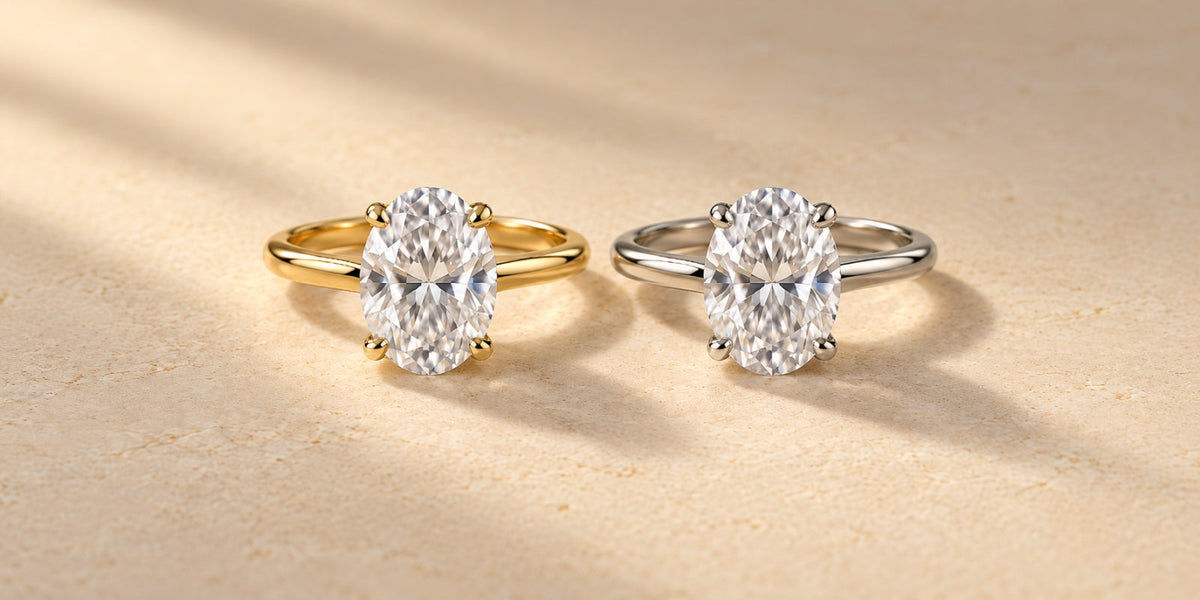 1. Matching oval diamond solitaire engagement rings in yellow gold and white gold displayed side by side on a warm ivory stone surface.