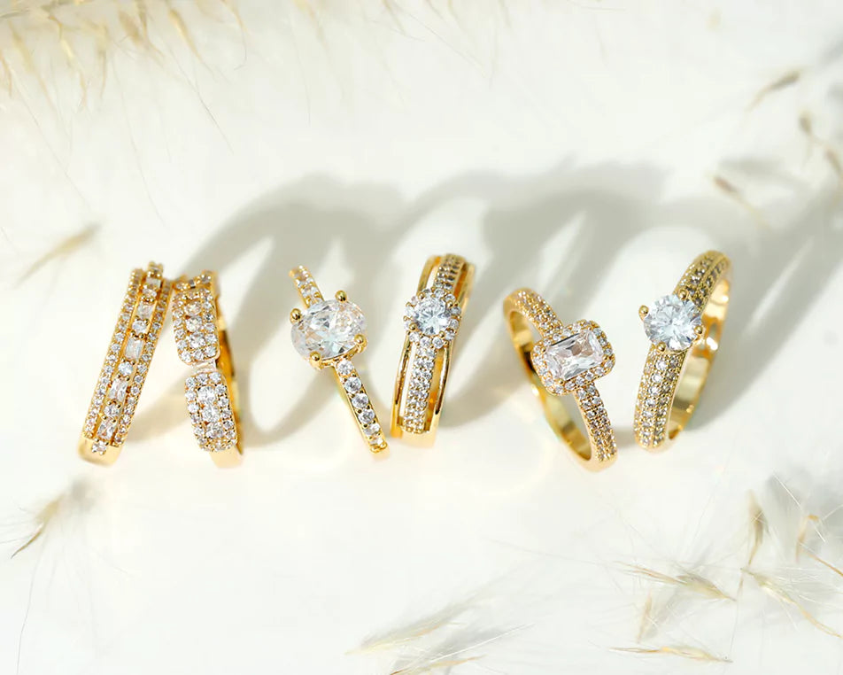 Collection of six luxurious gold lab-grown diamond rings displayed on a soft, neutral background with delicate feather-like accents. Each ring features a unique design: from double bands of pavé-set diamonds, to solitaire settings with oval, round, and emerald-cut center stones. The intricate gold bands are adorned with various styles of side stones, creating a cohesive yet diverse bridal or fashion ring set that sparkles under natural lighting.