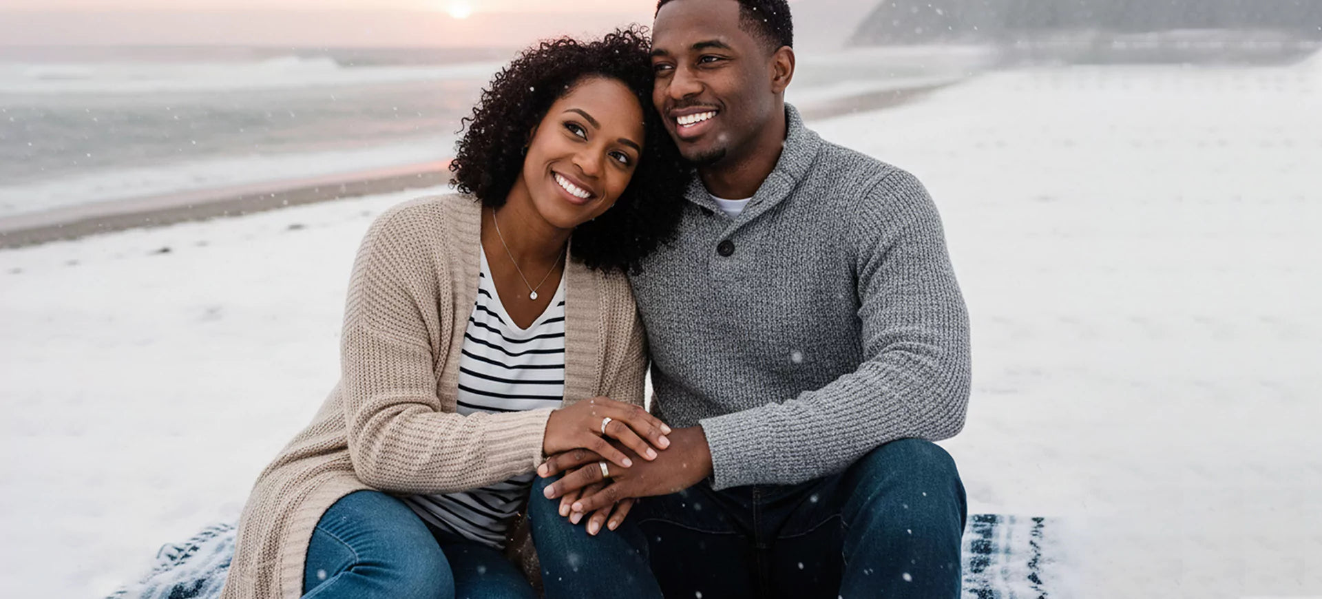  Smiling couple sitting by the snowy beach, showcasing elegant jewelry with her sparkling engagement ring and delicate pendant necklace glowing in natural light.